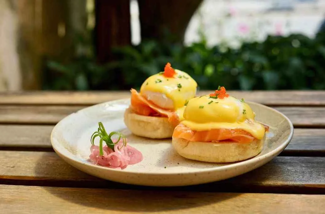 Trứng Benedict phủ sốt vàng trên bánh mì cùng thịt nguội trên đĩa sứ