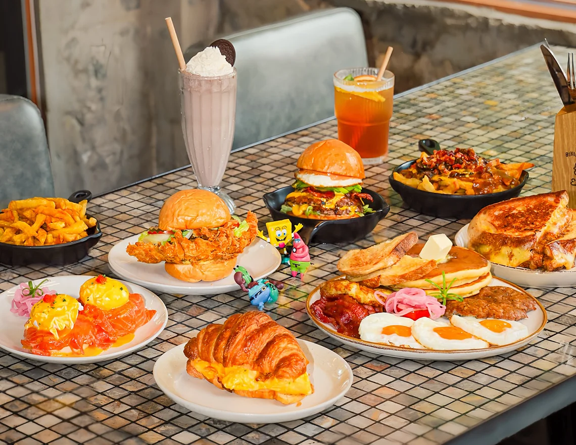 Bàn tiệc brunch nhiều món gồm burger, khoai tây chiên, bánh ngọt và đồ uống tại Bikini Bottom Express